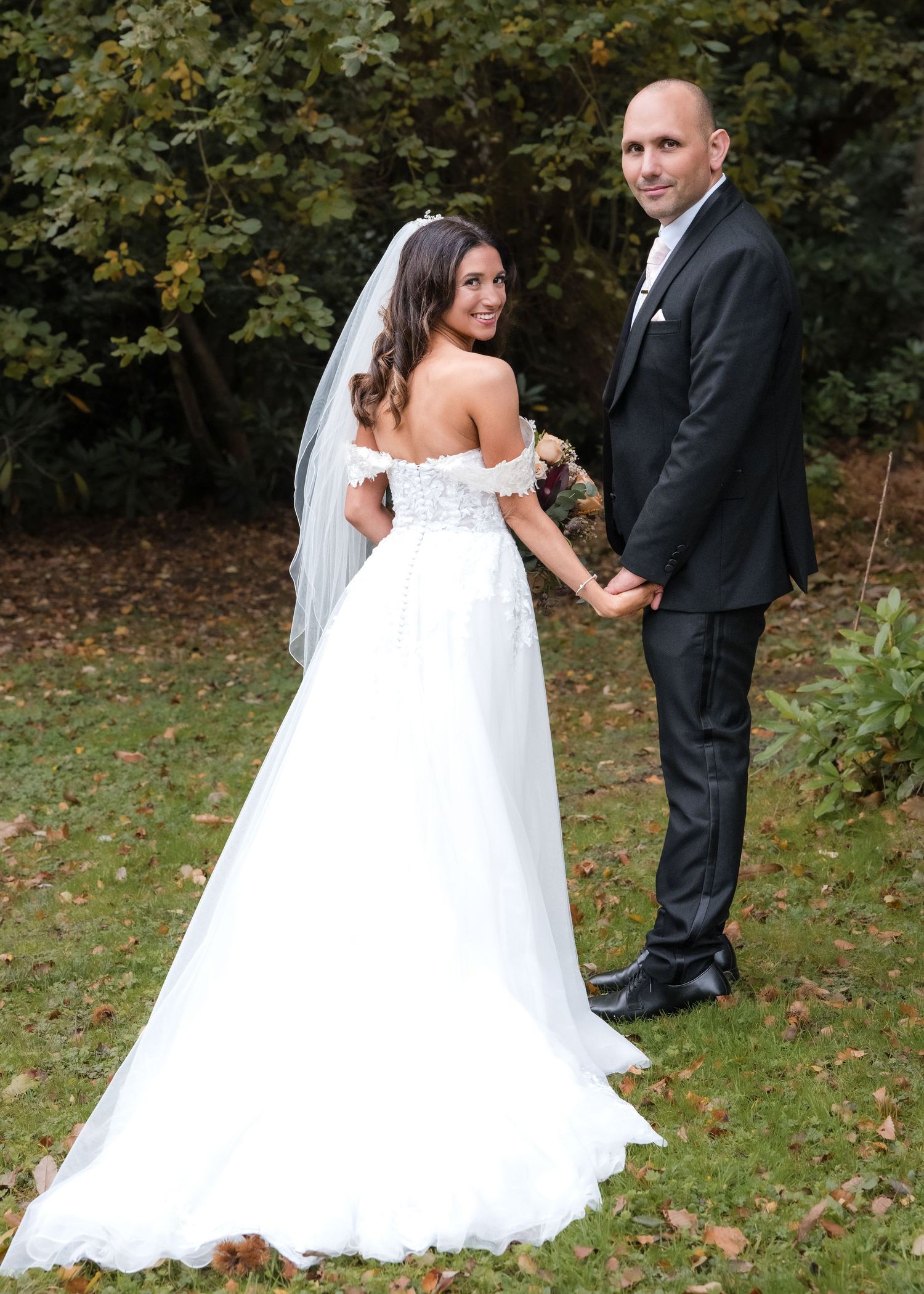 Bride in off-shoulder white gown and groom in dark suit posing in a lush garden setting.