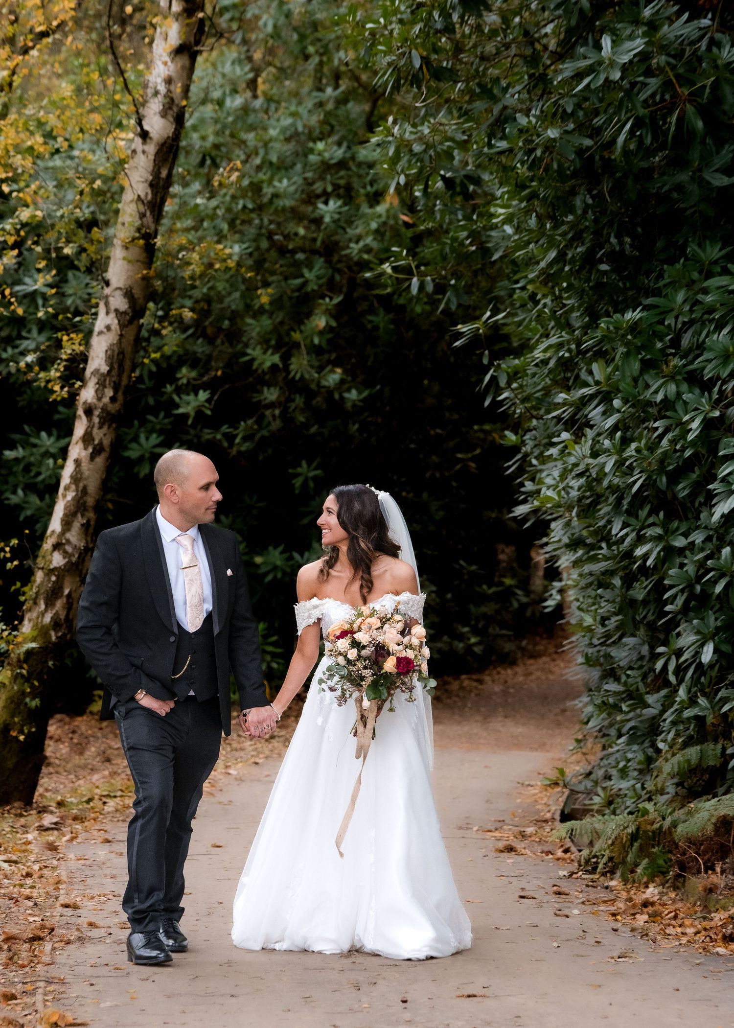 Bride and groom walking hand in hand along a tree-lined autumn path on their wedding day.