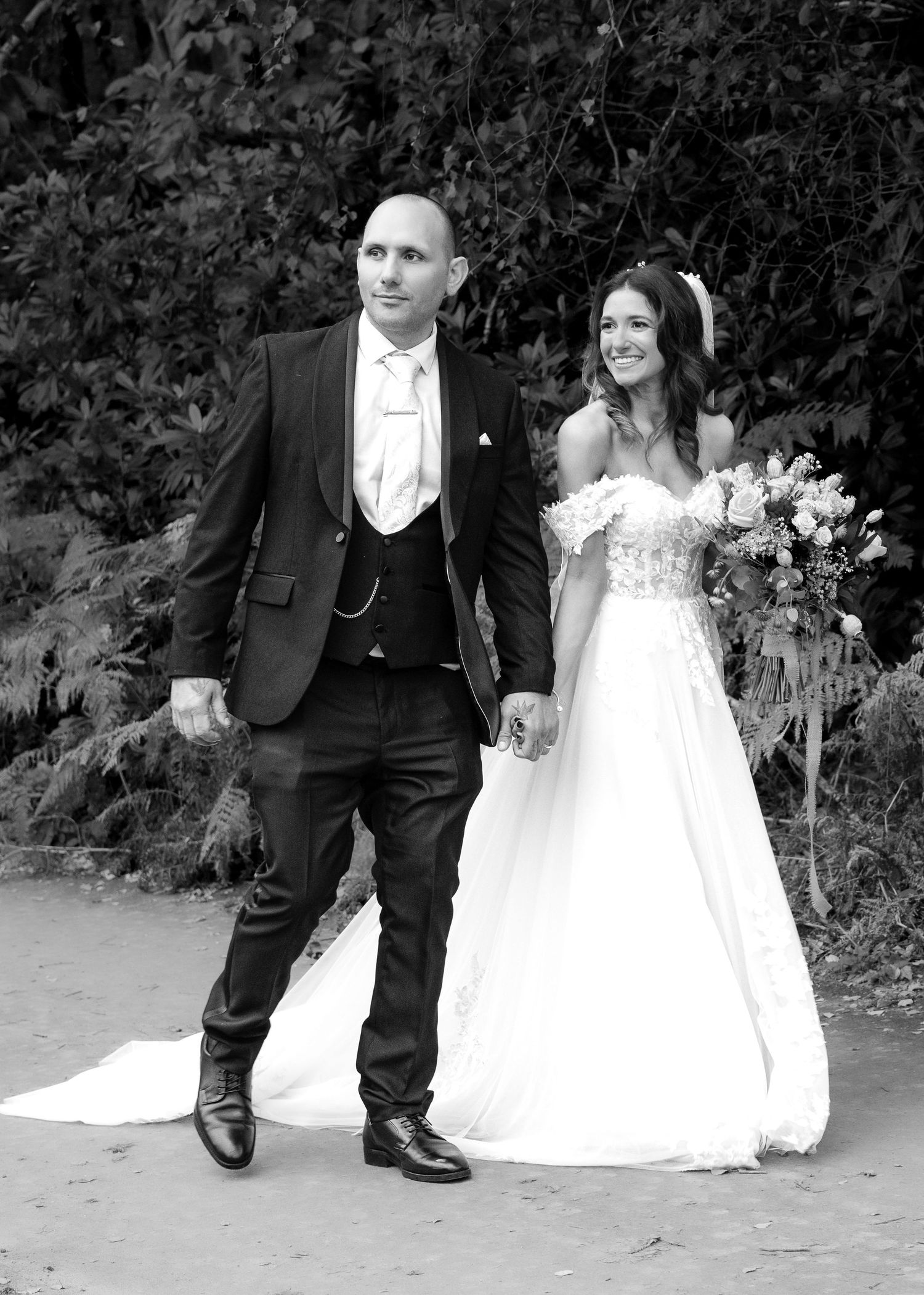 Black and white photo of bride and groom walking hand in hand along a garden pathway.