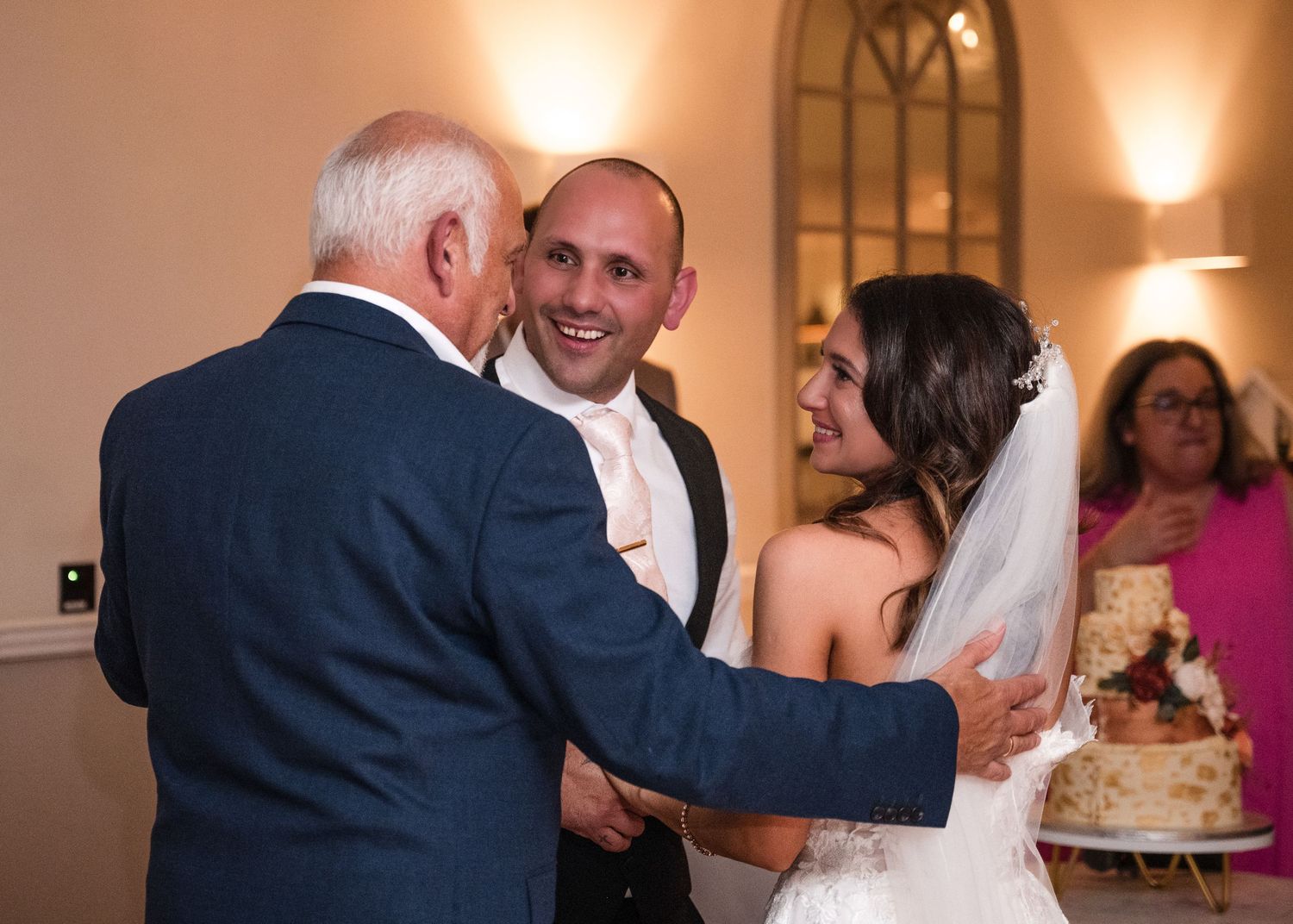 Smiling groom and bride greet an elderly guest near wedding cake at reception.