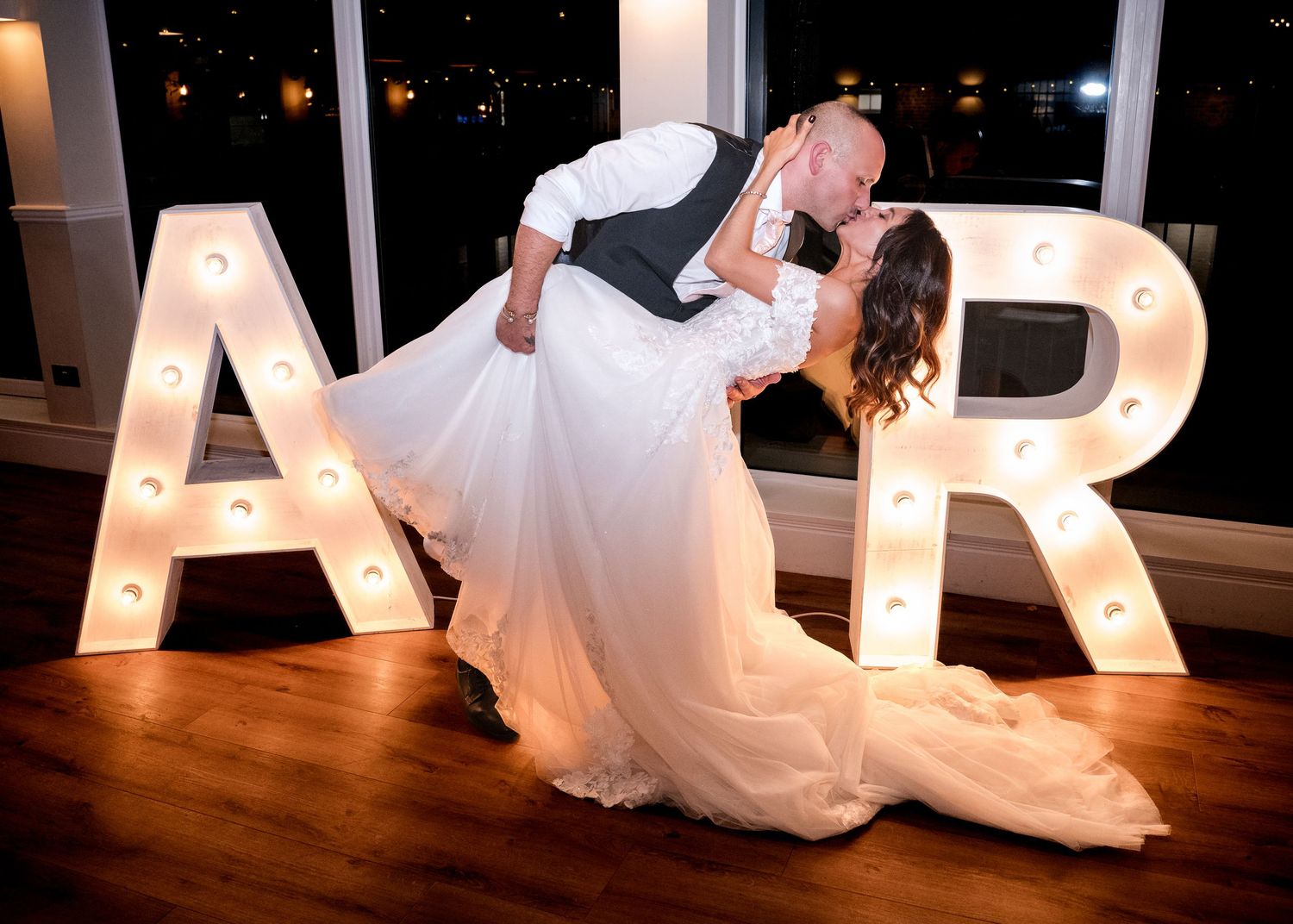 Bride and groom share a romantic dip kiss in front of illuminated marquee letters.
