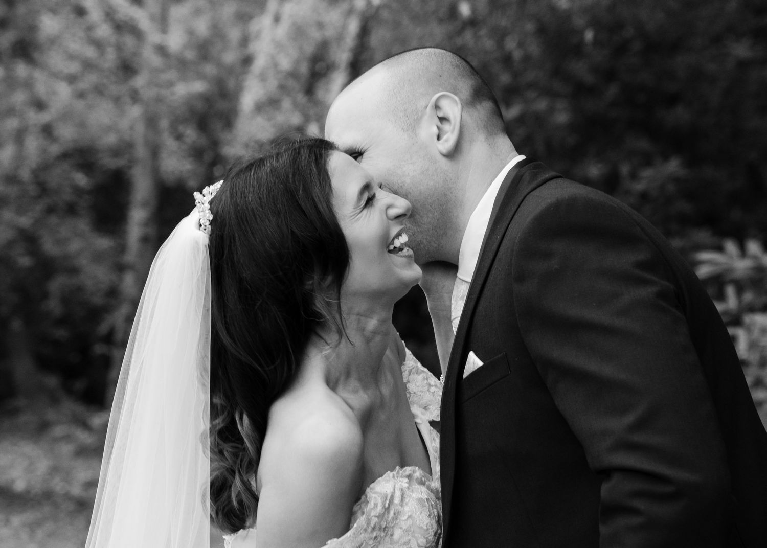 Black and white portrait of bride and groom smiling intimately in garden setting.