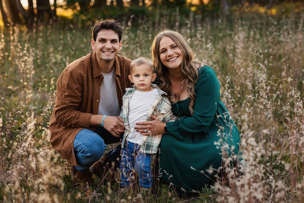 A smiling family of three poses in a golden field, with parents kneeling beside their toddler in warm autumn light.