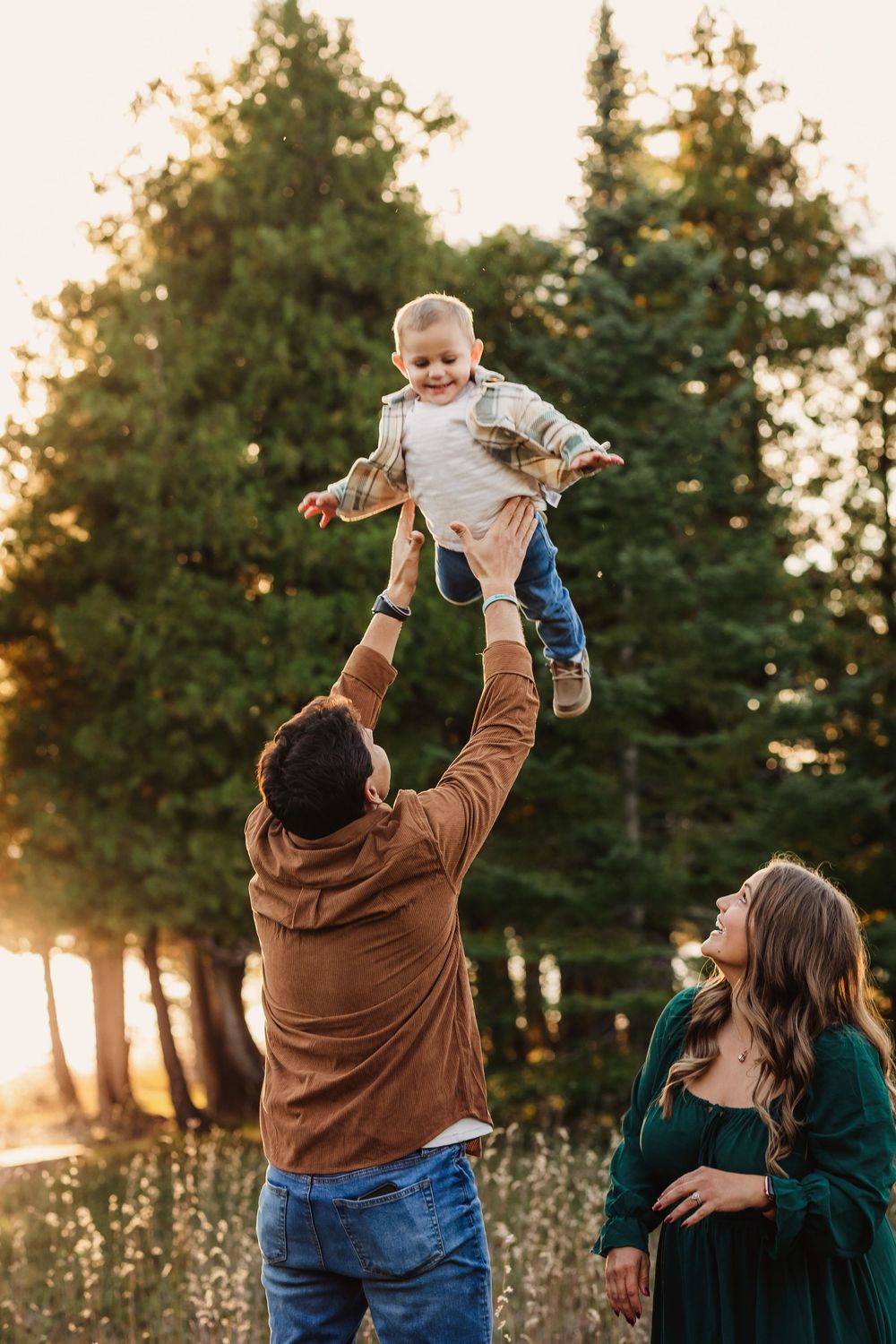 Father tossing smiling toddler in the air while pregnant mother watches, surrounded by pine trees in golden sunlight.