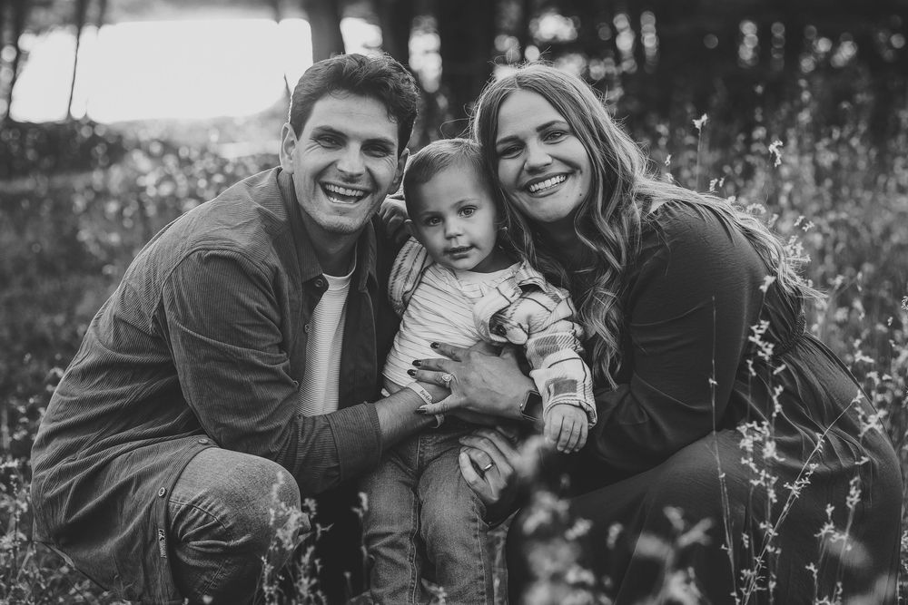 Black and white family portrait of smiling parents with toddler outdoors in a field.