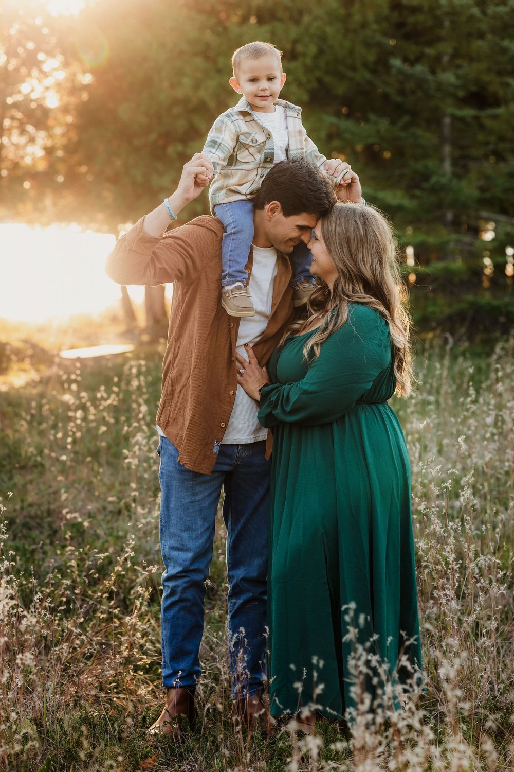 A family poses at golden hour in a meadow, parents touching foreheads while toddler sits on dad's shoulders.