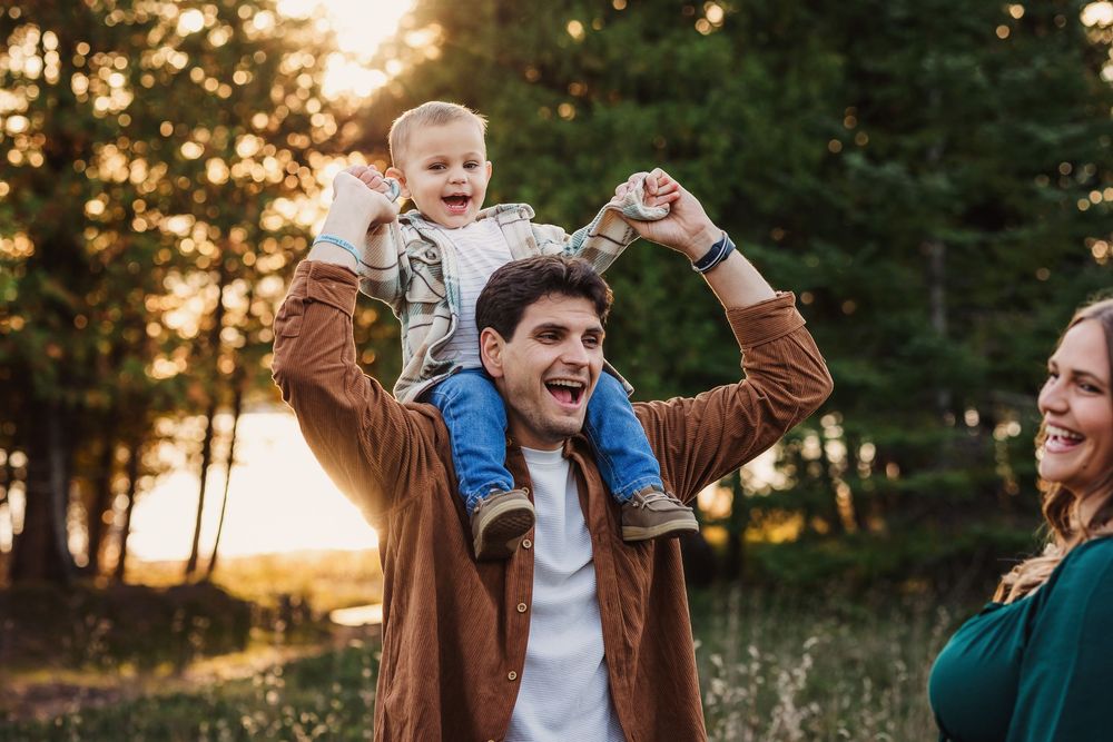 Happy family with father carrying laughing baby on shoulders and mother smiling outdoors at golden hour.