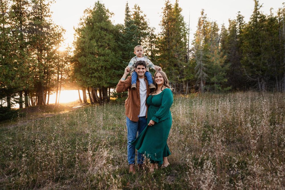 Family of three posing at sunset in a forest meadow, parents smiling with toddler on dad's shoulders.
