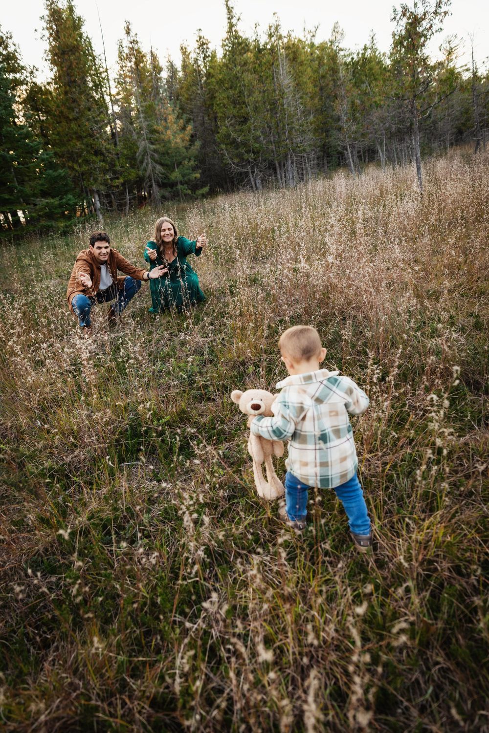 A toddler holding a stuffed animal runs toward smiling parents crouching in a golden meadow surrounded by tall trees.