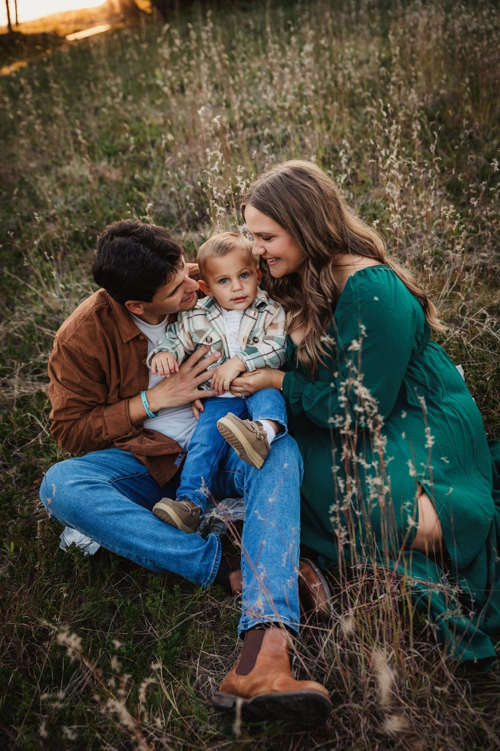 A family sits in a golden field at sunset, parents lovingly kissing their toddler son on the cheeks.