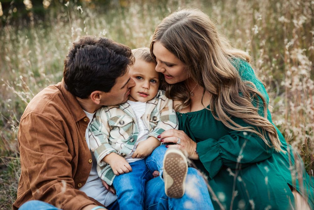 A happy family of three sits in a field, parents kissing their young son on the cheeks outdoors.