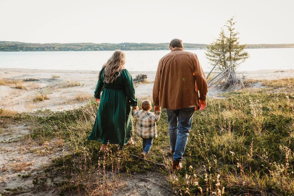 Family of three walking hand-in-hand toward a scenic lakeside landscape surrounded by wildflowers and trees.