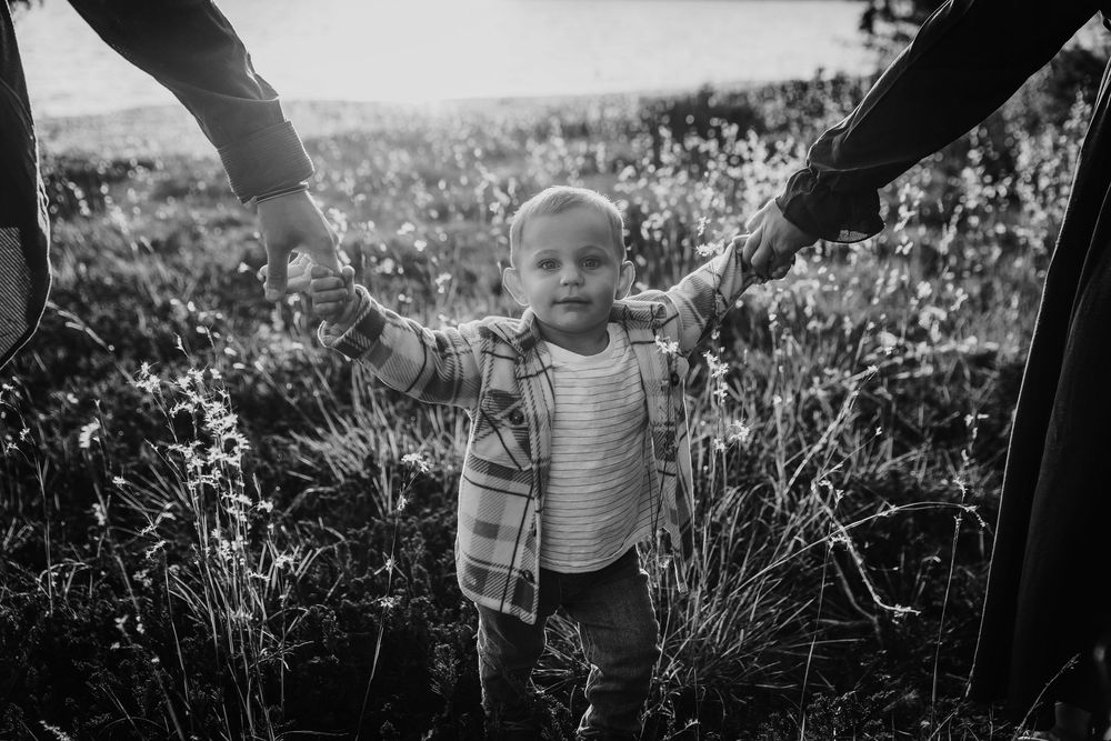 Black and white photo of a toddler holding hands with parents while standing in a wildflower field.