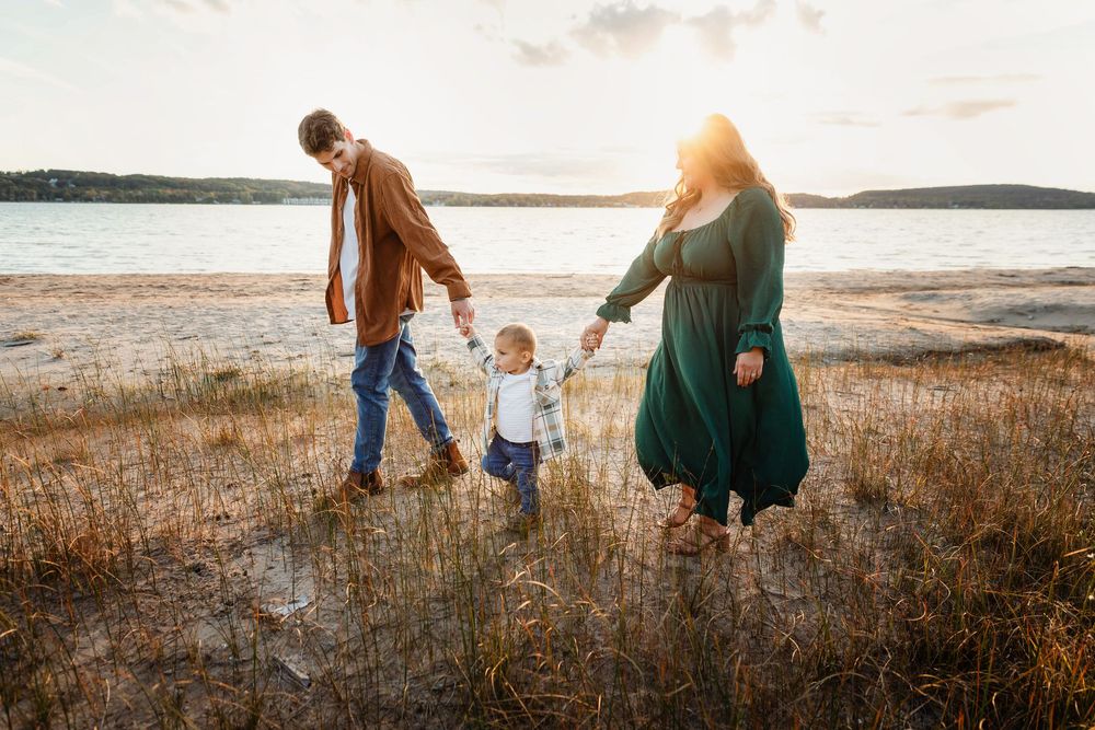 Family of three walking hand-in-hand along a sunlit sandy beach shore at golden hour sunset.