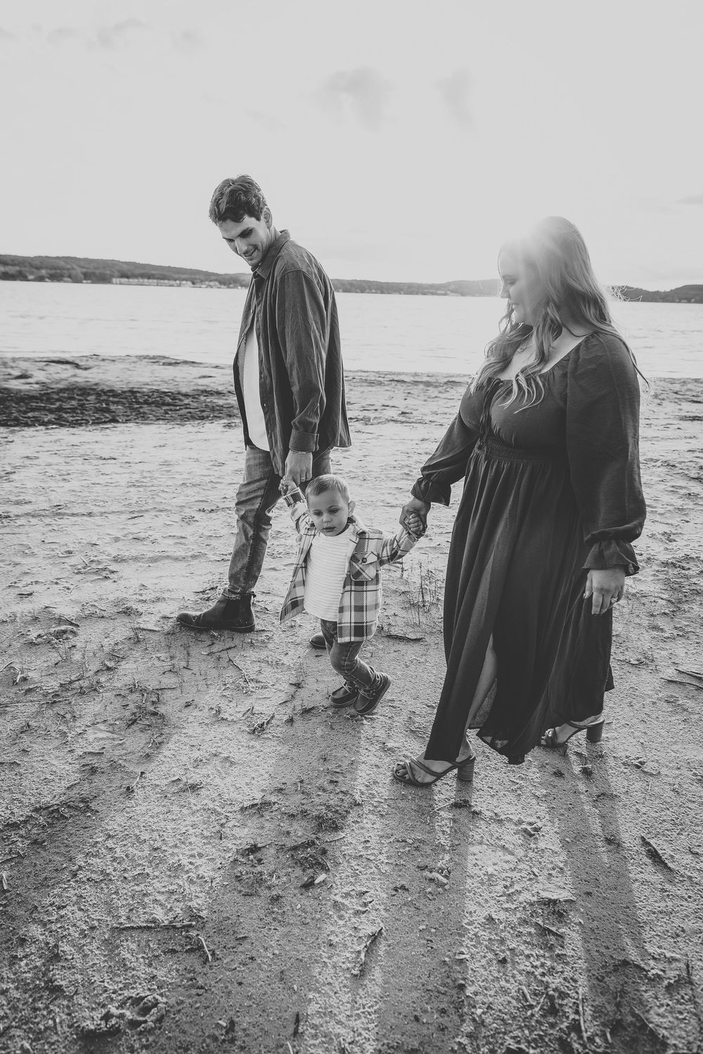 Black and white photo of a young family walking on a beach, parents holding hands with their toddler child.