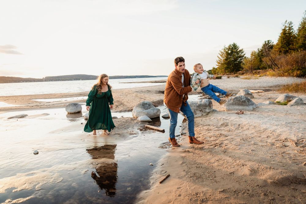 Family playing on a sandy beach at sunset, mother in green dress, father swinging toddler, trees and water in background.