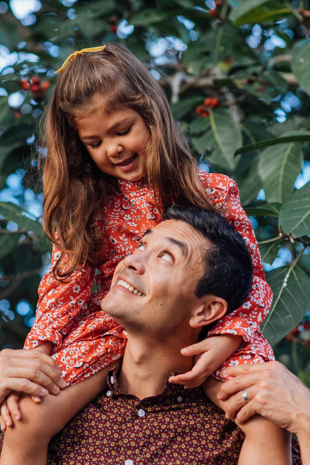 Happy father smiling up at young daughter in red floral dress sitting on his shoulders outdoors.