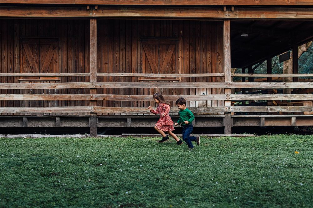 Two young children in colorful outfits running and playing on green grass near wooden building.