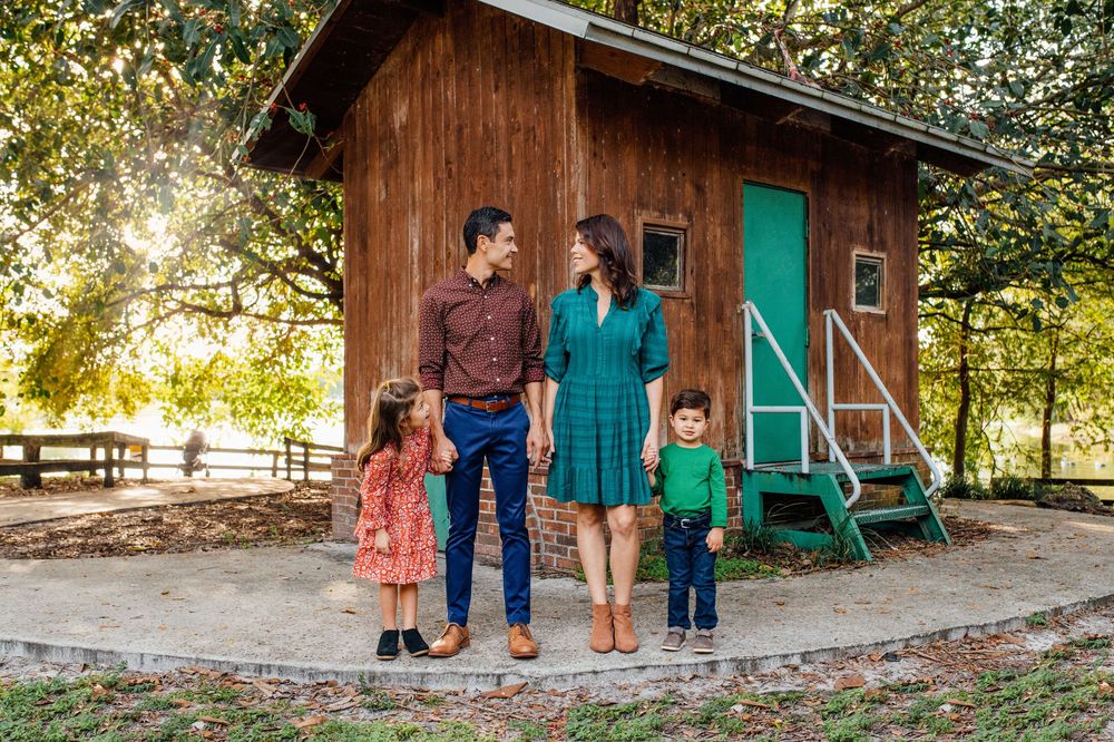 Happy family of four standing in front of rustic wooden cabin surrounded by trees outdoors.