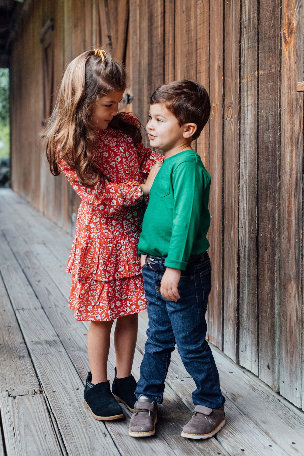 Young girl in red floral dress standing with younger boy in green shirt on wooden porch outdoors.