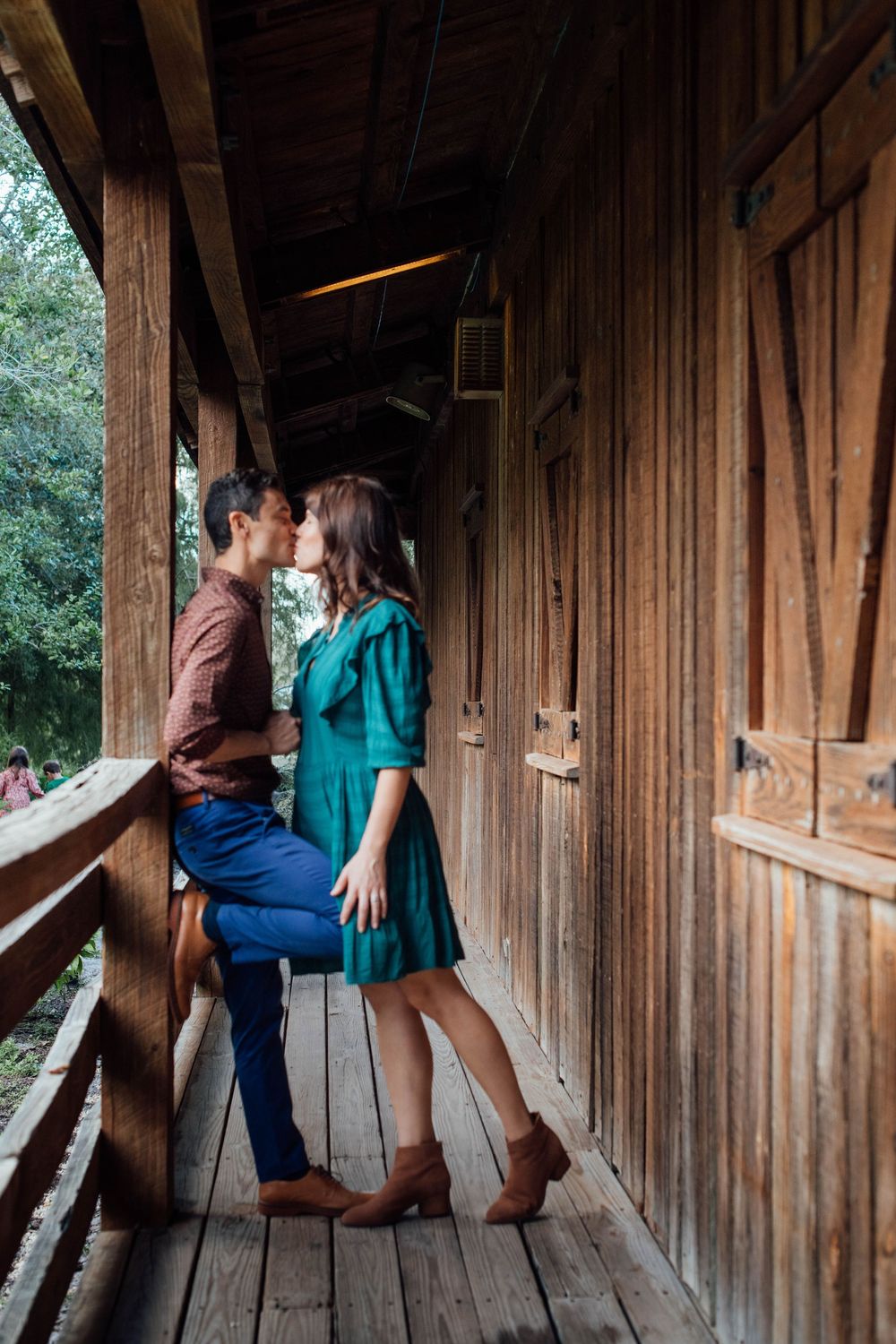 Couple sharing a romantic kiss on a rustic wooden porch balcony surrounded by lush green trees.