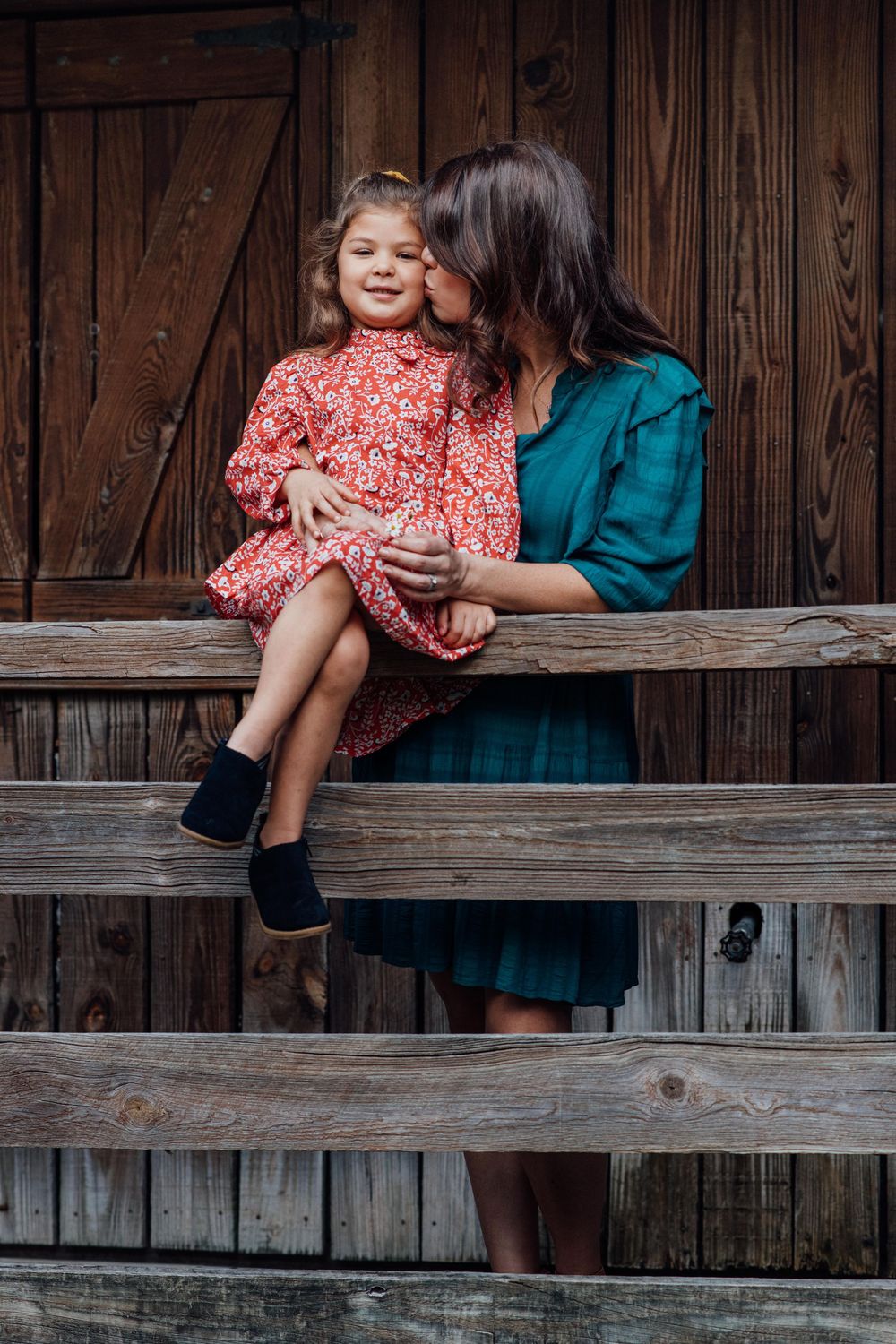 Mother in teal dress kissing young daughter in red floral dress on wooden steps of rustic barn.