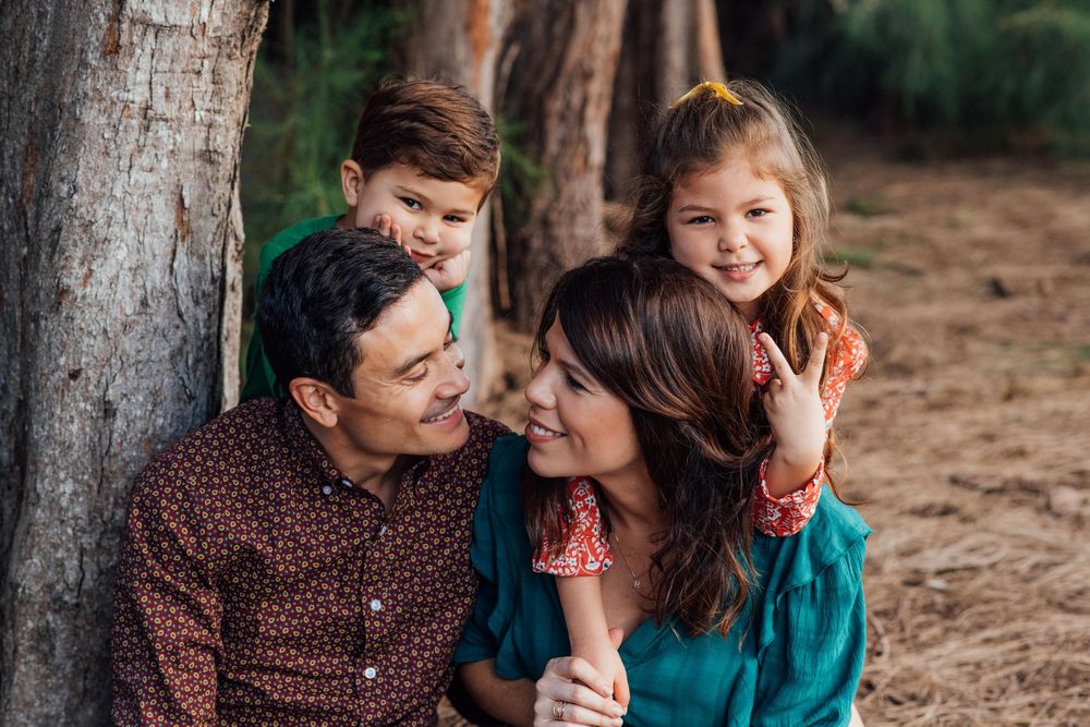 Joyful family of four laughing together beside a tree trunk in a warm outdoor setting.