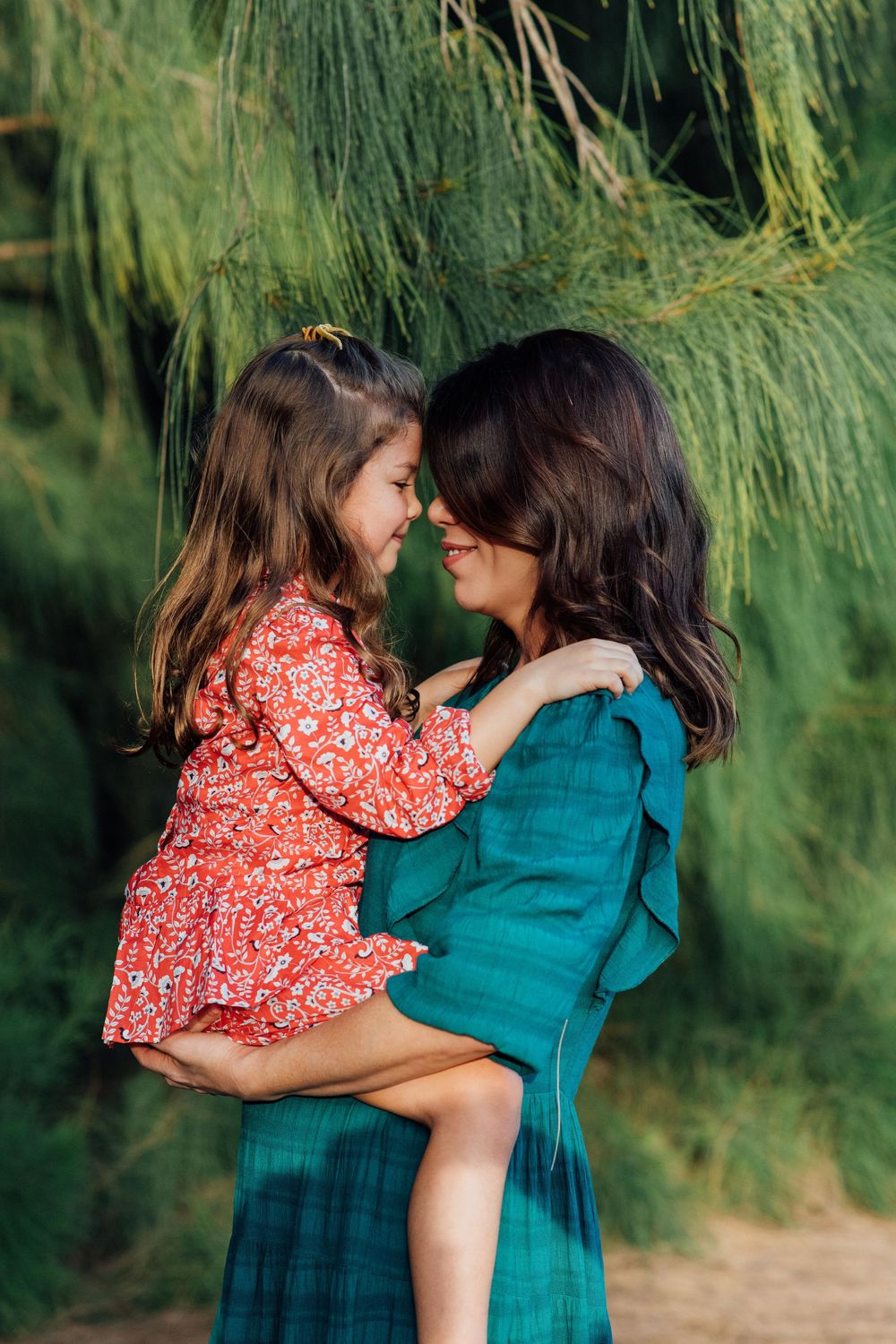 Mother in teal dress holding young daughter in red floral dress against lush green pine branches.