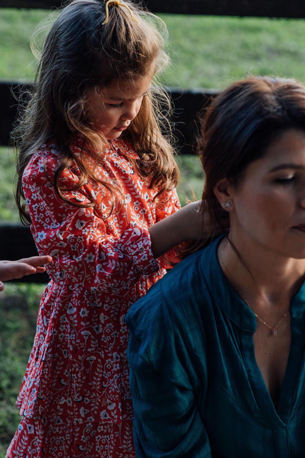Young girl in red floral dress touching mother's shoulder in a warm golden outdoor light.