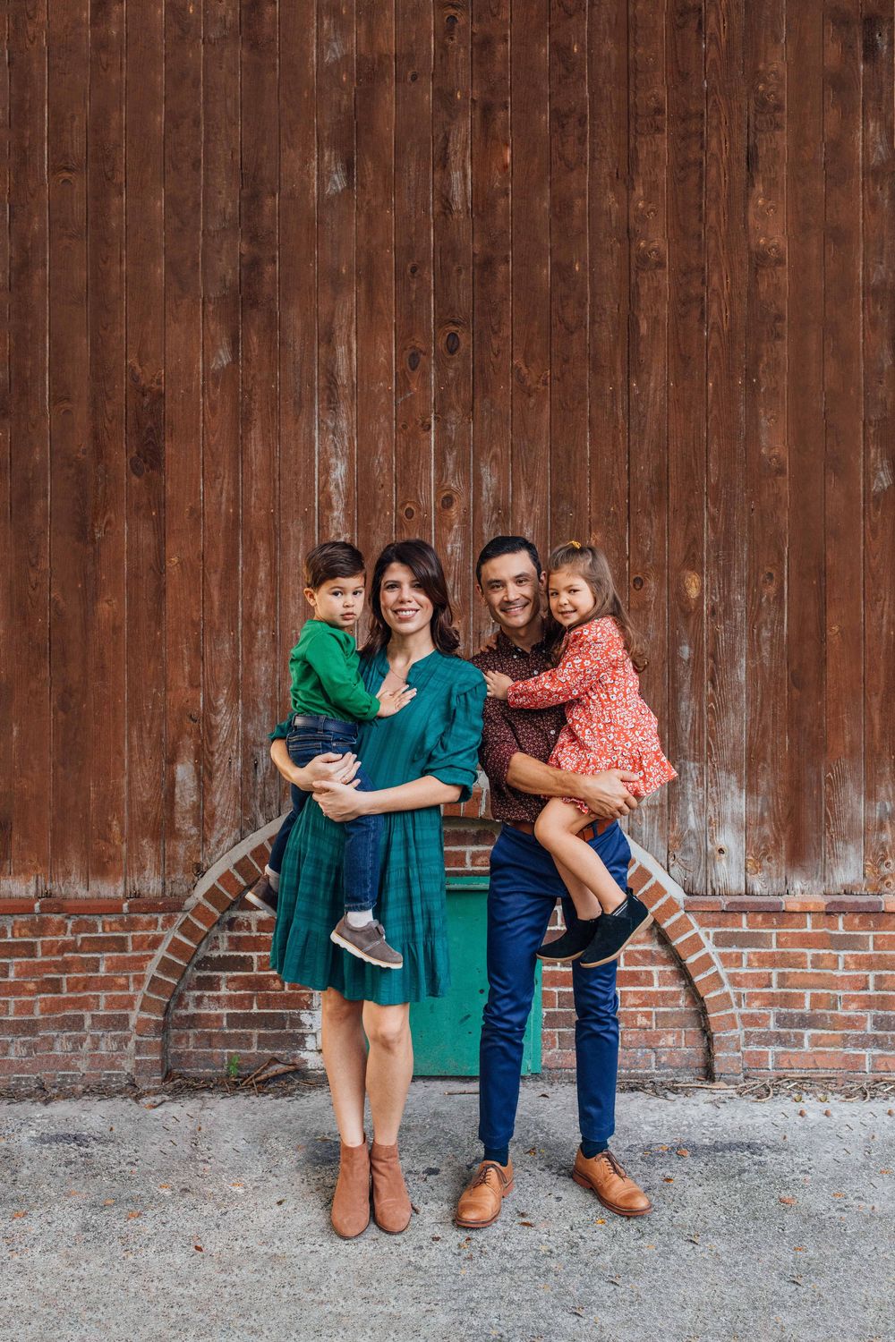 Smiling family of four posing together in front of rustic wooden barn wall, parents holding children.