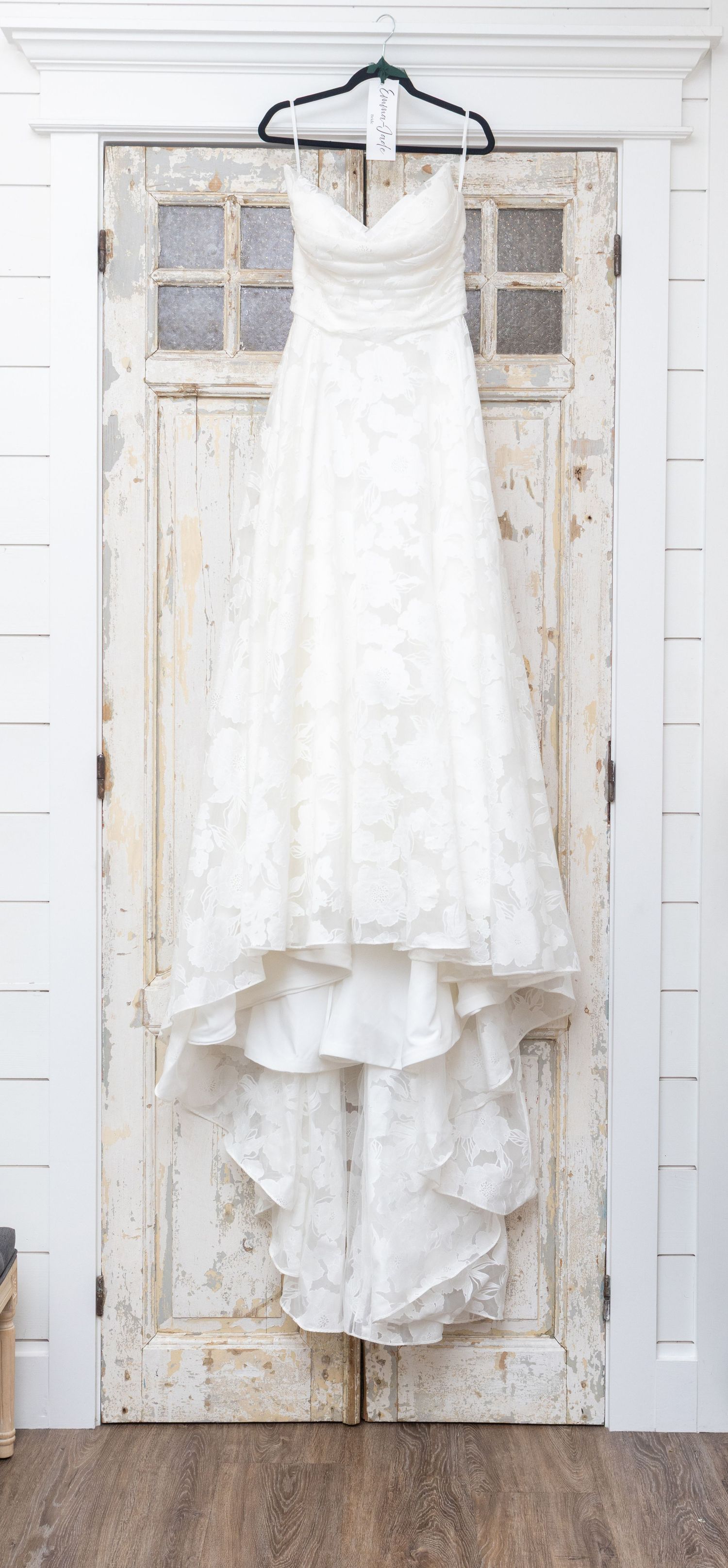 White strapless A-line wedding dress with floral lace detail hanging on a rustic distressed white door.