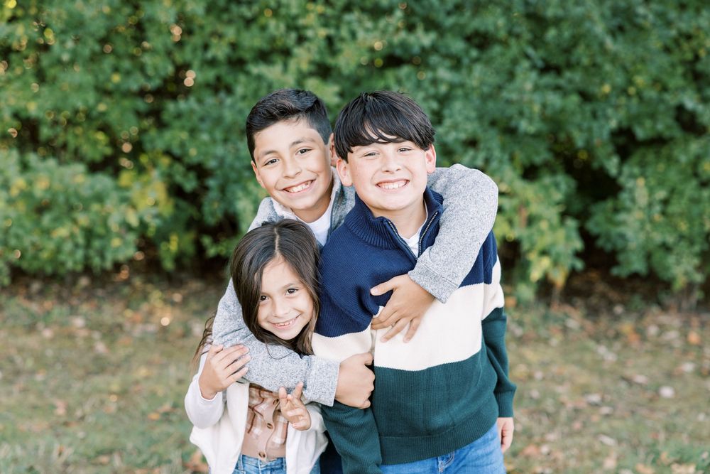 Three smiling siblings hugging outdoors in front of lush green bushes.