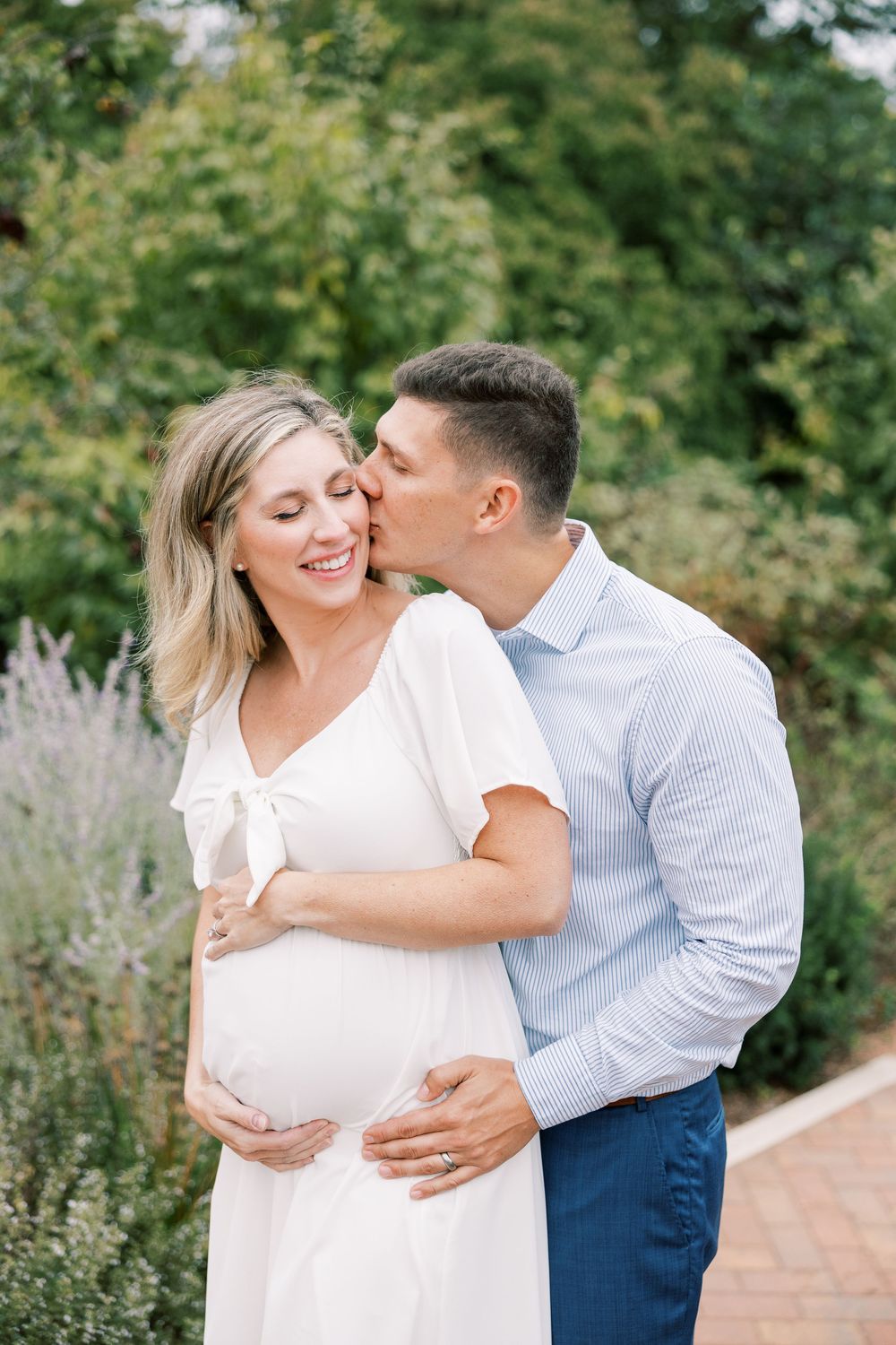 Pregnant woman in white dress smiling as her partner kisses her cheek in a garden setting.