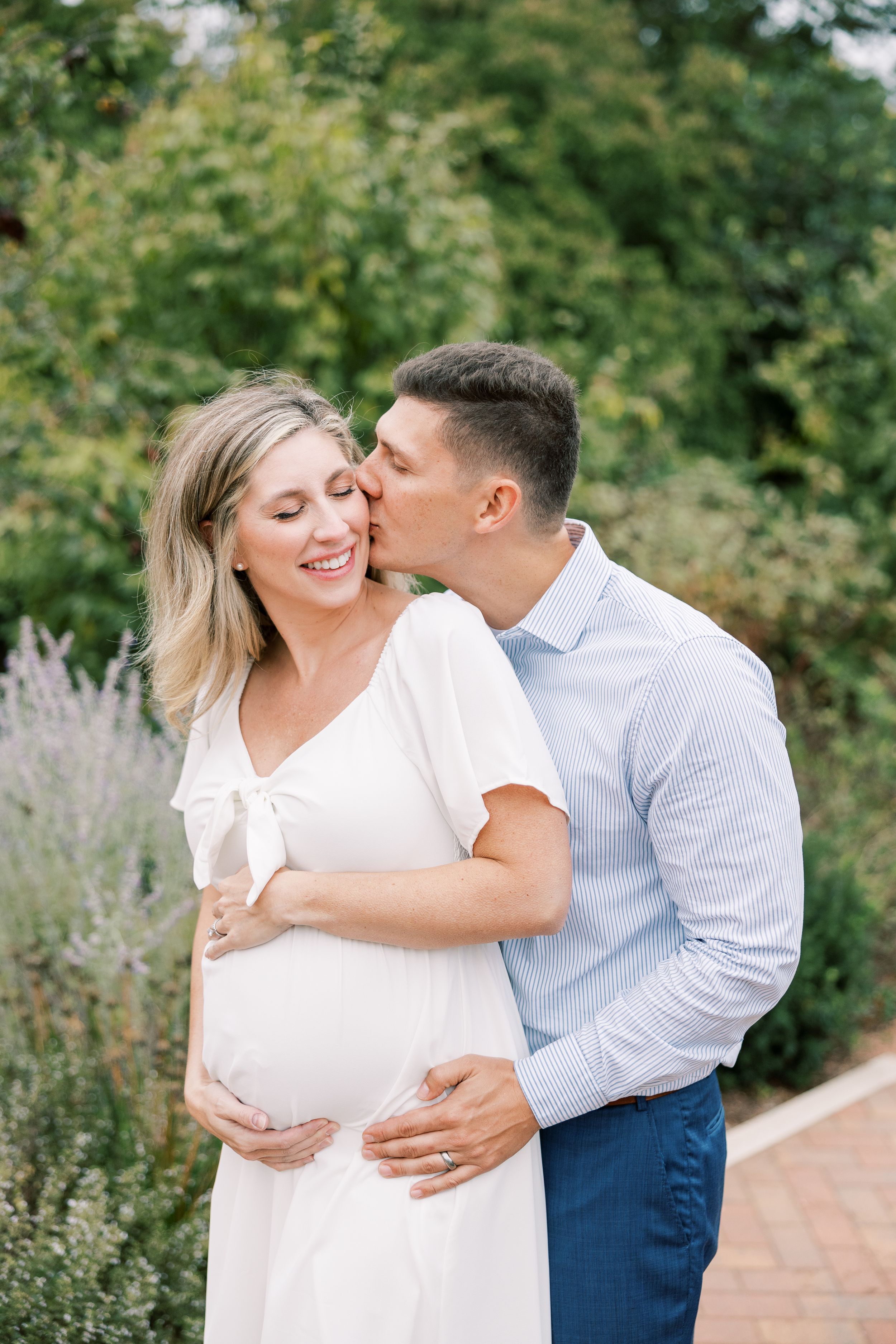 Pregnant woman in white dress smiling as her partner kisses her cheek in a garden setting.