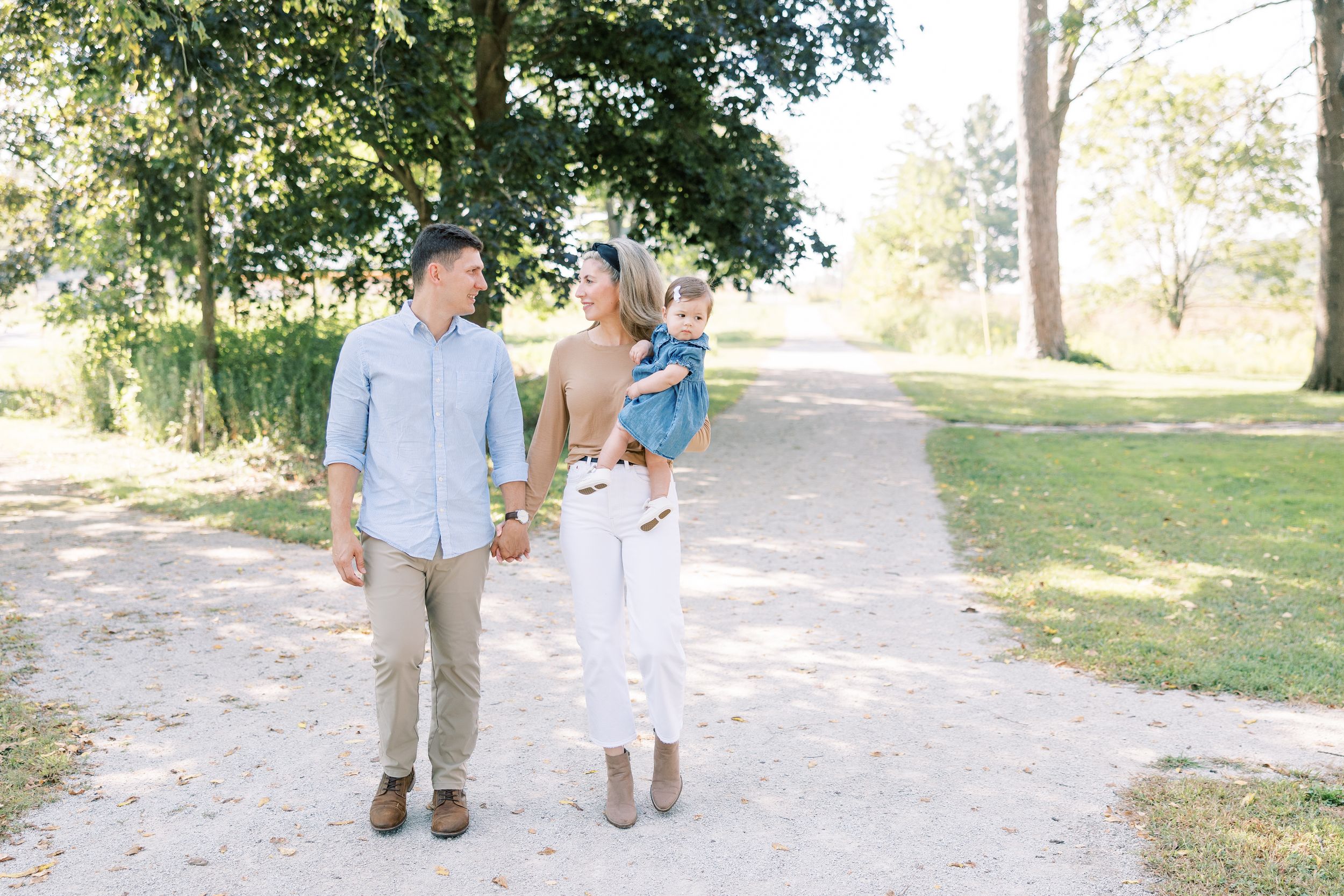 Young family with toddler walking hand-in-hand on a sunny park path surrounded by green trees.