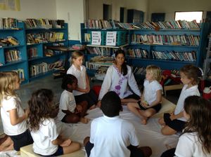 Teacher leads a group of children in a circle discussion on the floor of a school library with blue bookshelves.