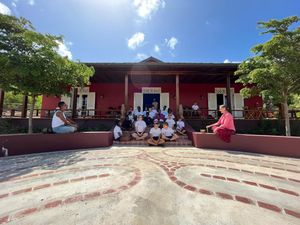 Group of students sitting outside a vibrant red colonial building with trees and blue sky in a tropical setting.
