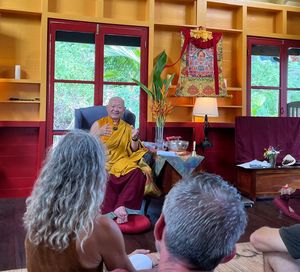 Tibetan Buddhist monk in yellow robes teaching students in a decorated meditation hall with thangka artwork.