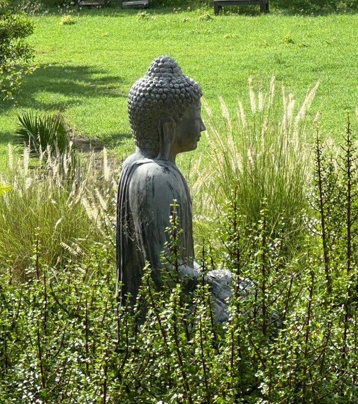 A serene Buddha statue meditating among lush green ornamental grasses and wildflowers in a peaceful garden setting.
