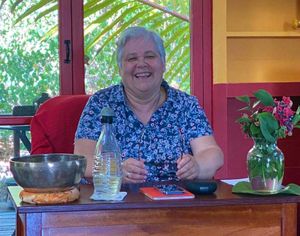 Smiling woman sitting at wooden table with water bottle, singing bowl, and pink flowers in bright tropical setting.