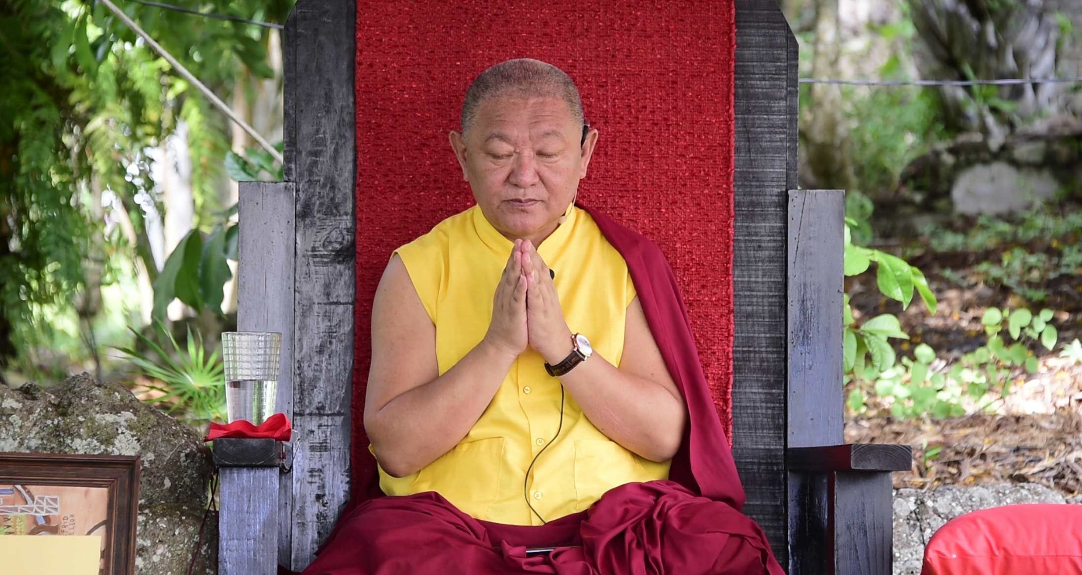 Tibetan Buddhist monk in yellow and red robes meditating with hands in prayer position outdoors.
