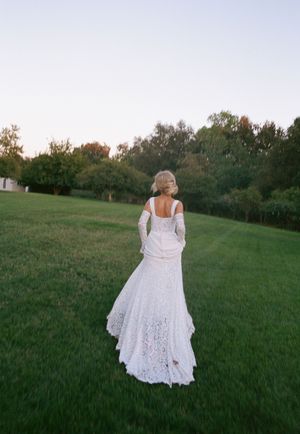 A person in a flowing white lace wedding dress walks through a serene green field at sunset.