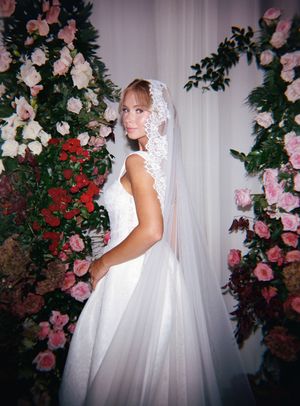 A bride in a flowing white wedding gown and veil poses among pink and red floral arrangements against a white backdrop.