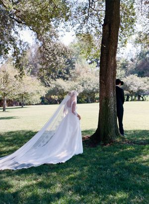 A flowing white wedding dress with a long train billows gracefully in the breeze against a backdrop of trees in a park setting.