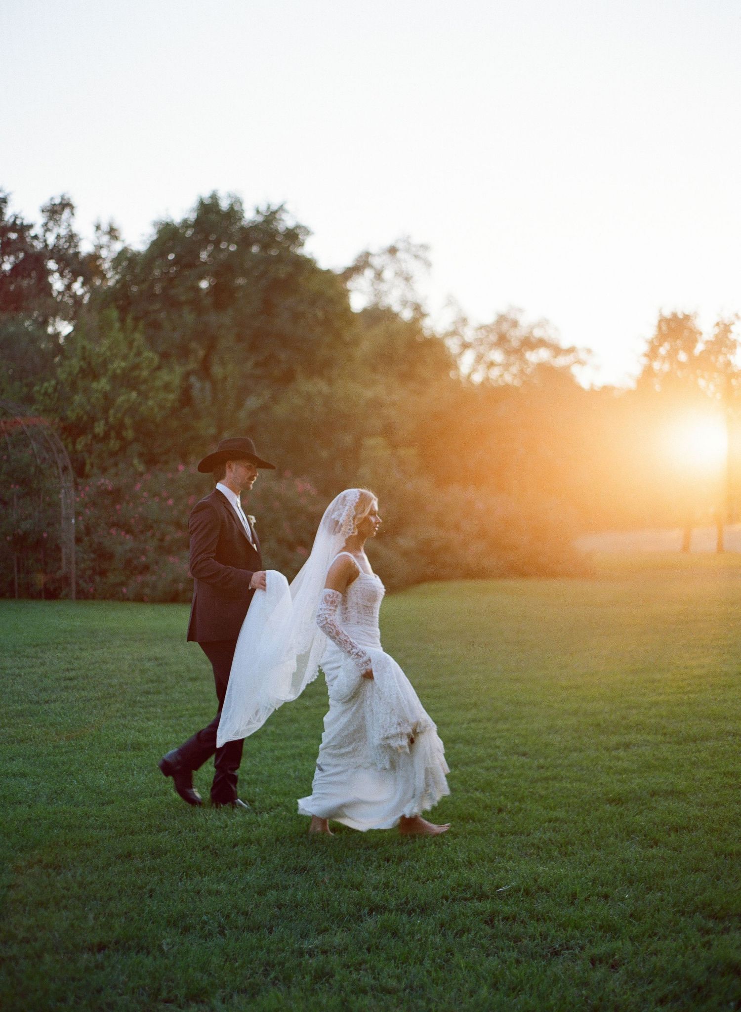 A couple in wedding attire runs playfully across a grassy field at sunset creating a romantic silhouette.