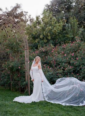 A bride in a flowing white wedding dress stands in a garden setting as her long veil catches the breeze at sunset.