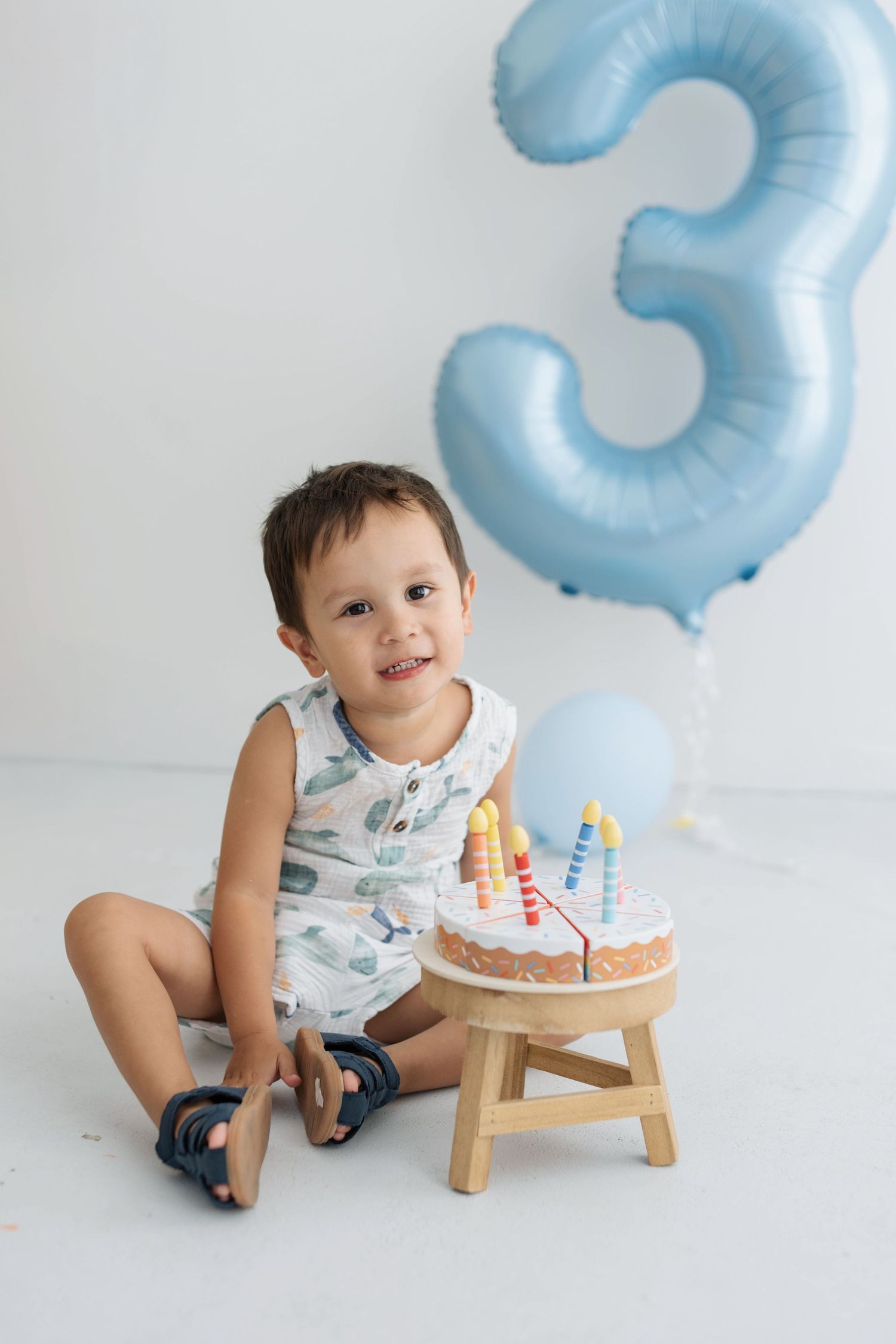 Smiling toddler boy sitting by birthday cake with candles, celebrating 3rd birthday with blue number balloon.