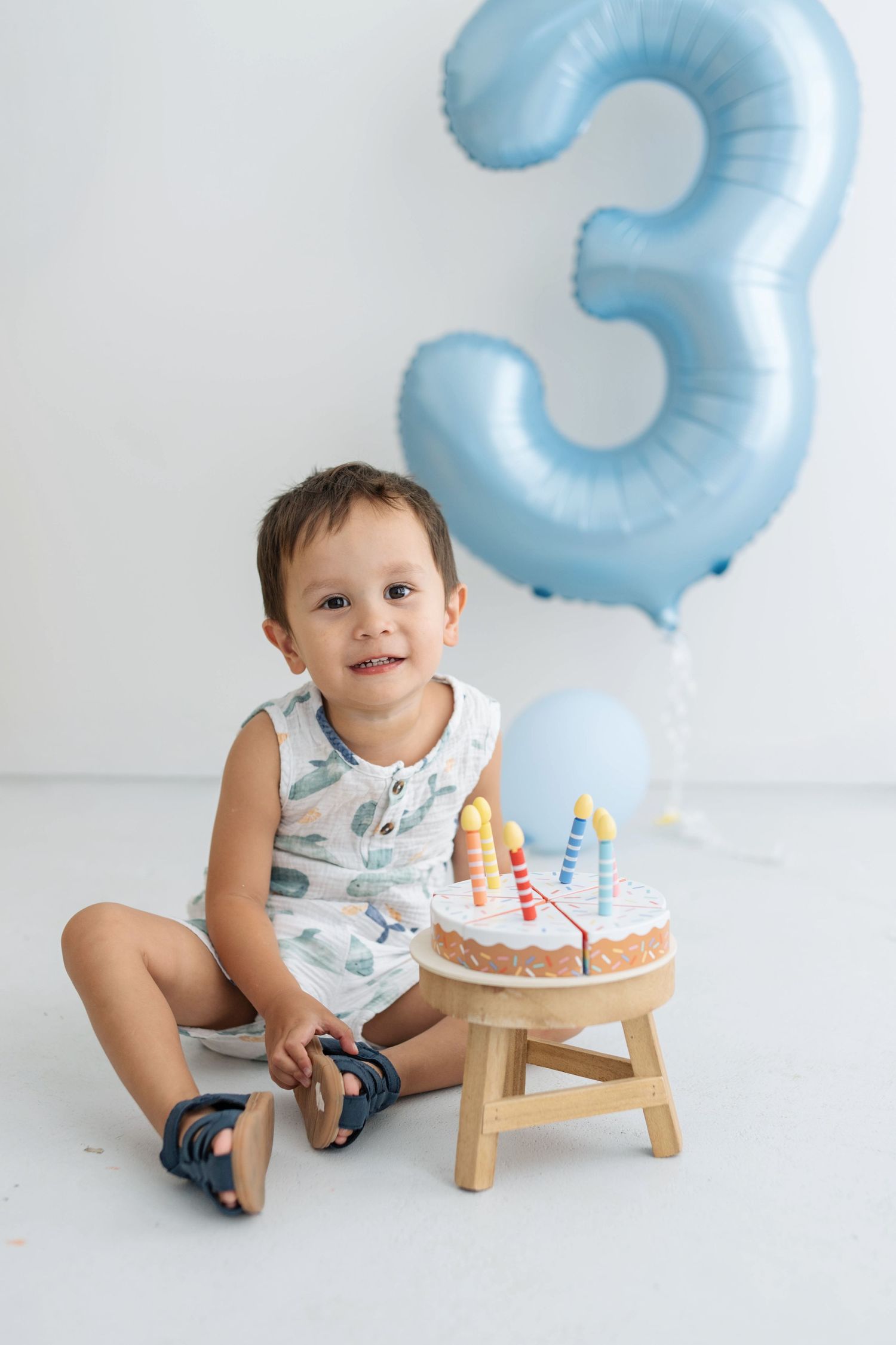 Smiling toddler boy sitting beside birthday cake with candles, celebrating his 3rd birthday with blue balloon backdrop.