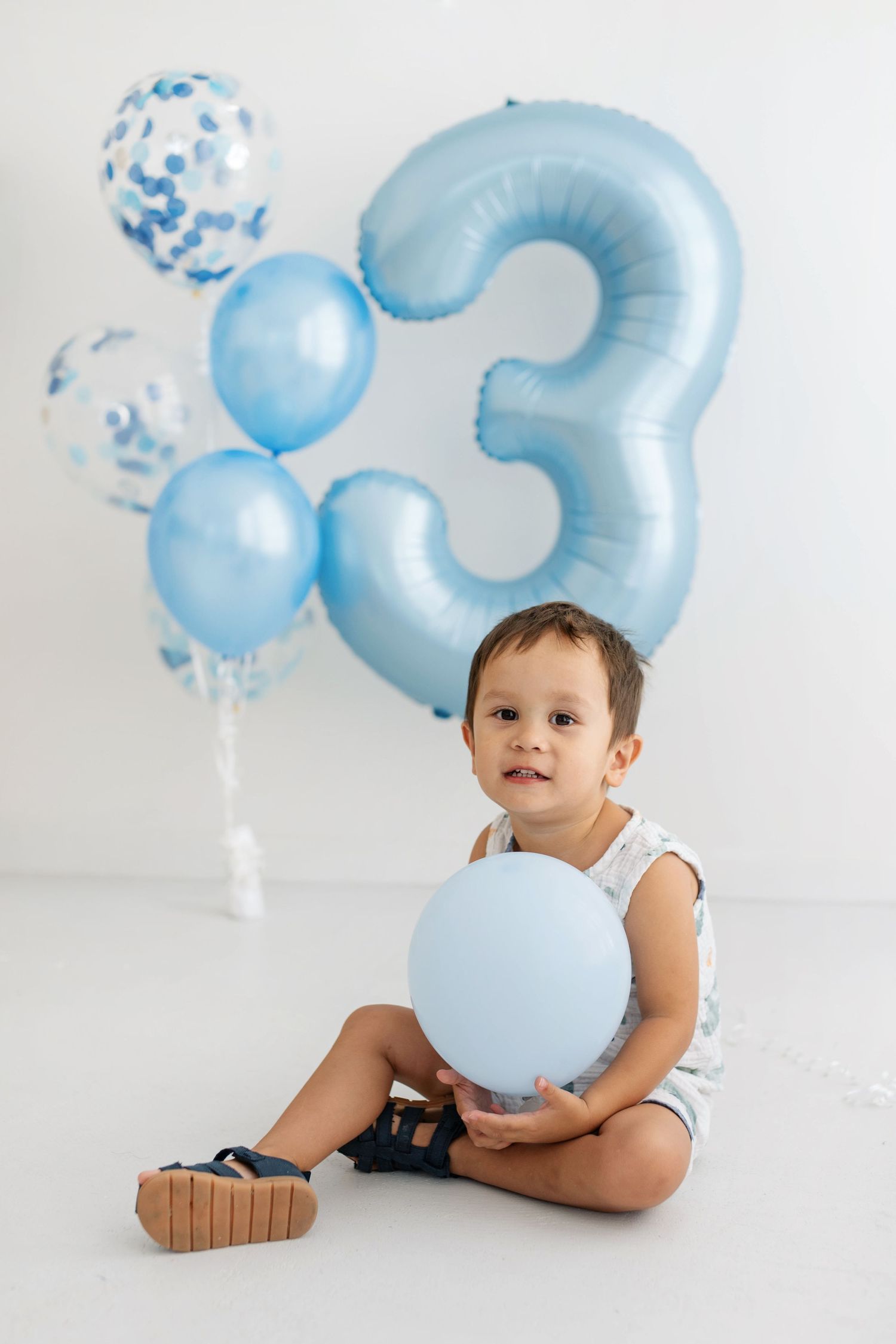 A toddler boy sits on a white floor holding a blue balloon, celebrating his 3rd birthday with blue number and confetti balloons.