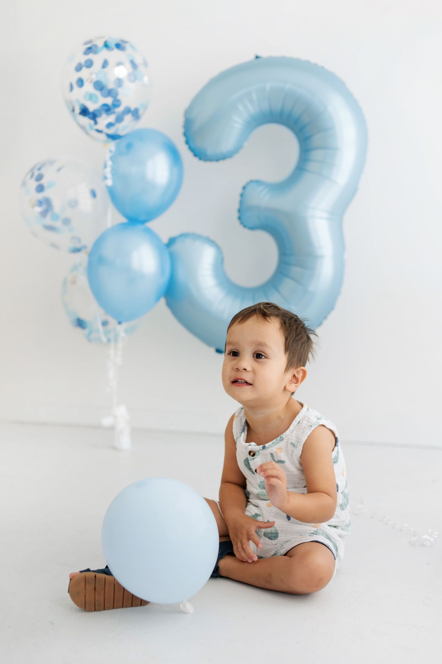 Toddler boy sitting on white floor with blue balloons and number 3 balloon celebrating his 3rd birthday.